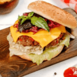 A gourmet burger with a sesame seed bun, a juicy beef patty topped with melted cheddar cheese, fresh lettuce, a dollop of creamy sauce, vibrant red cabbage slaw, and garnished with basil leaves, served on a wooden board.